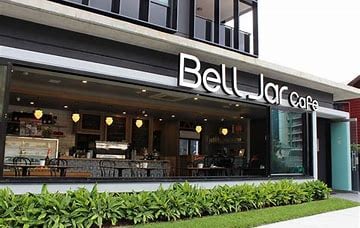 The exterior of Bell Jar Cafe showcases modern architecture with large glass windows displaying the interior. Green landscaping lines the entrance, enhancing the inviting atmosphere. The cafe sign is prominently displayed, with warm lighting illuminating the space, suggesting a comfortable spot for guests to enjoy. 