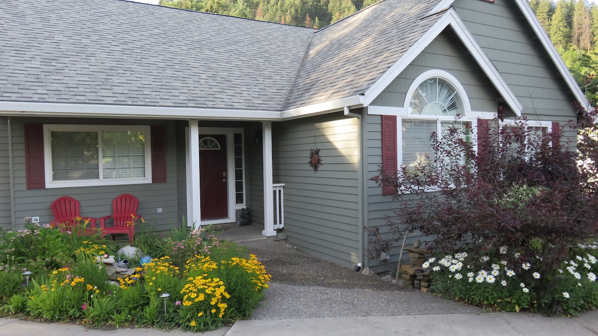 The entrance of a remodeled home is framed by colorful flower beds, featuring yellow and white blooms. A pathway of pebbles leads to the front door, which is complemented by two red Adirondack chairs. Lush greenery surrounds the area, enhancing the welcoming appearance.