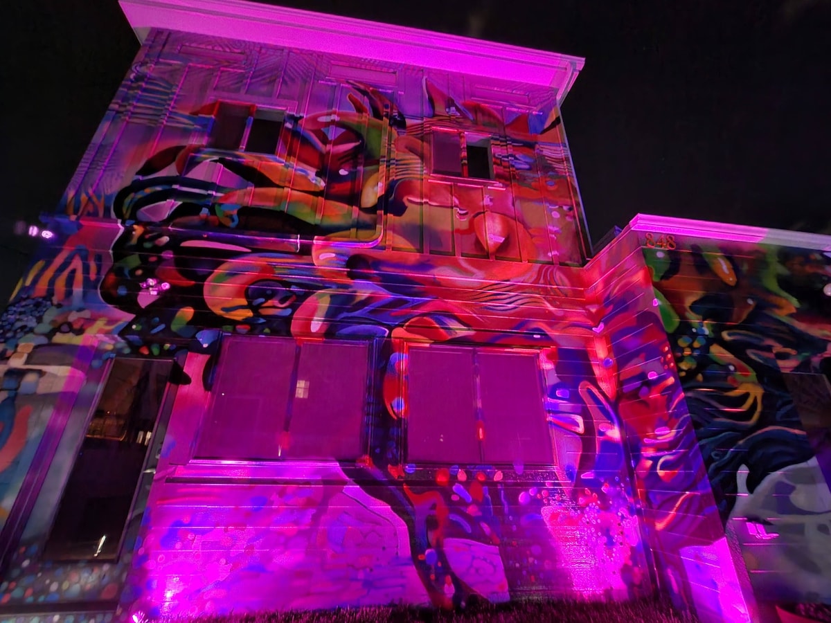 The exterior wall of the property is adorned with a vibrant mural, illuminated in shades of pink and purple. The artwork features abstract shapes and fluid patterns, creating a dynamic visual effect at night, enhancing the property's colorful aesthetic.