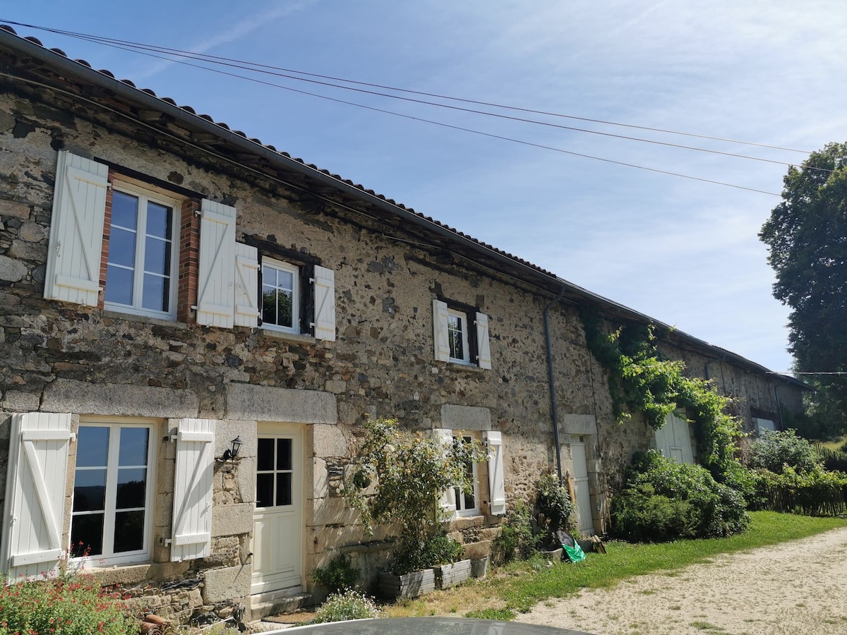 The exterior of a rustic stone house is presented, featuring white shutters and multiple large windows. Lush greenery and flowering plants surround the entrance, inviting a sense of tranquility. A clear blue sky complements the charming appearance of the home.