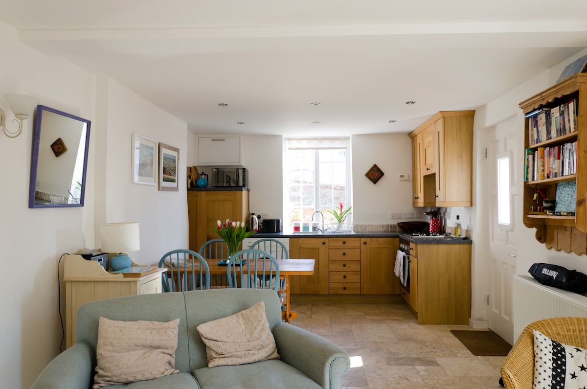 An open-plan kitchen and sitting area is displayed, with a comfortable two-seater sofa facing a wooden dining table for four. The kitchen features modern appliances, including a microwave and oven, while natural light fills the space through a window above the sink.