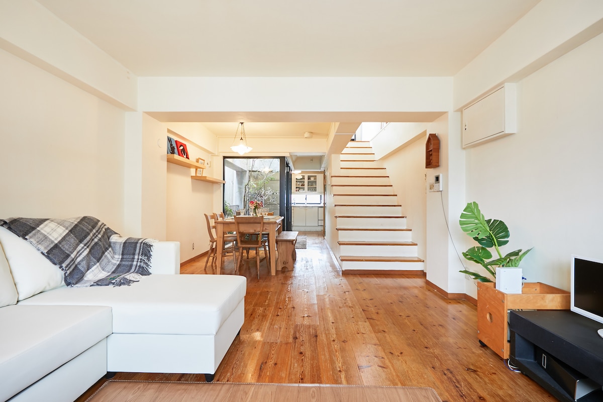 A spacious living area features warm wooden floors, complemented by a white sectional sofa adorned with a patterned throw. A dining table set for six is positioned nearby, with a large window allowing natural light to illuminate the space. A staircase leads to the upper floor.