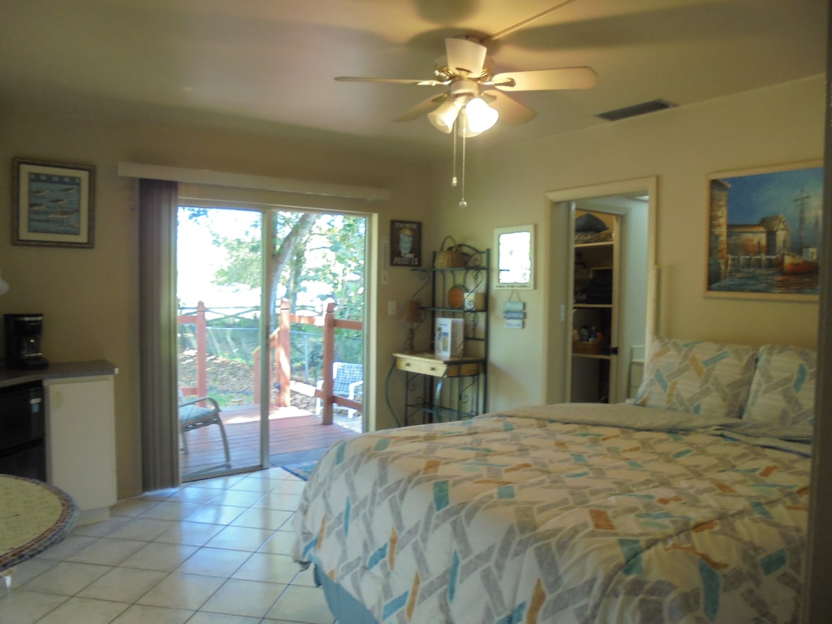 A cozy bedroom is equipped with a spacious bed featuring a patterned duvet cover. A ceiling fan offers airflow, while sliding doors lead to a private deck. A small kitchenette is visible, including a coffee maker and a seating area. Natural light illuminates the room.
