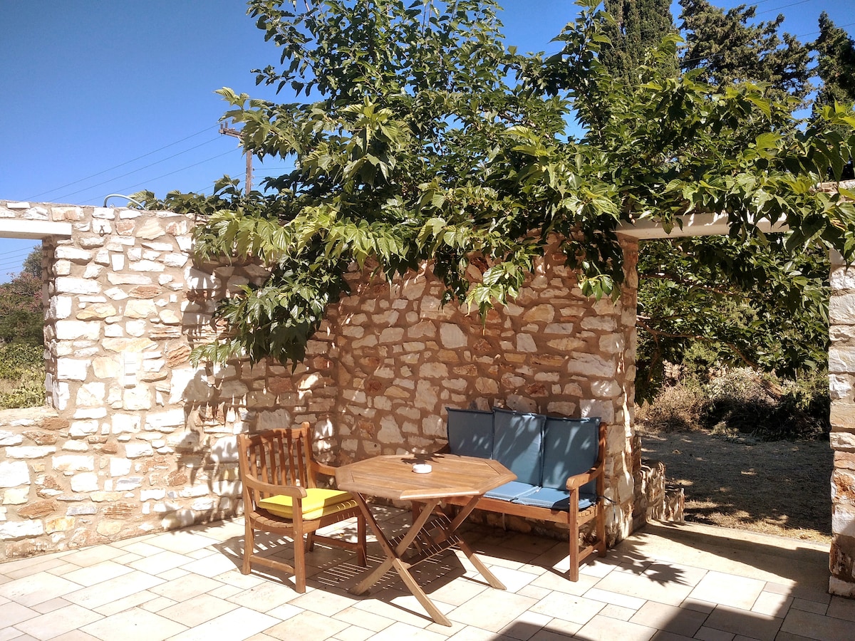 An outdoor seating area features a round wooden table surrounded by a wooden bench and a chair. Natural stone walls provide shade, while green foliage adds a refreshing touch. The tiled patio area creates a welcoming space for relaxation.