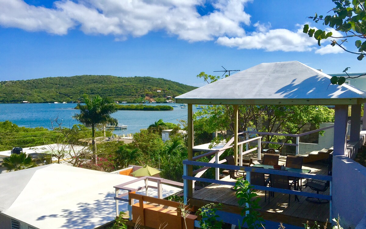 The image captures a scenic view of Ensenada Honda, showcasing a waterway bordered by lush greenery and hills in the background. A shaded deck with outdoor seating is visible, inviting relaxation while enjoying the natural beauty. Bright blue skies and fluffy clouds enhance the serene atmosphere.