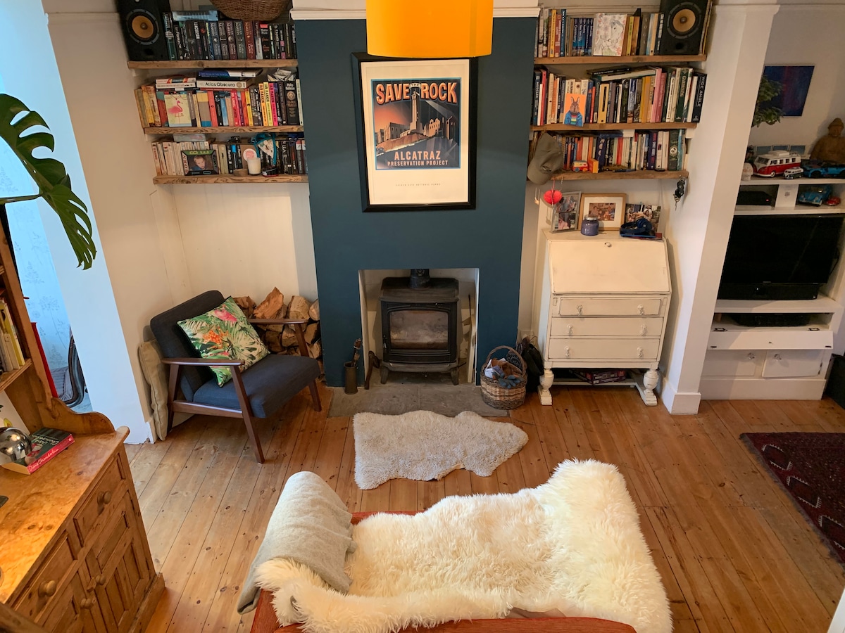 A cozy living area features a comfortable armchair and a patterned rug over warm wood flooring. A fireplace is positioned against a dark accent wall, complemented by shelves filled with books and decorative items. Soft light is introduced through a hanging pendant lamp.