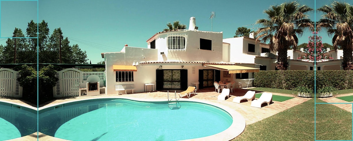 The villa features a private pool surrounded by well-maintained gardens. Lounge chairs are positioned beside the pool, offering a relaxing space. A barbecue area is visible under the shaded patio, providing a convenient space for outdoor dining.