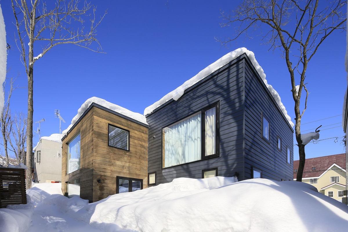 A modern townhouse is depicted, showcasing two distinct sections with contrasting wood and dark cladding. The roof is covered with snow, while a snowy landscape surrounds the building. Clear blue skies enhance the winter scene, providing a serene backdrop.