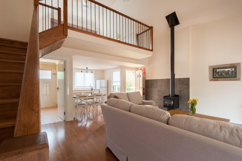 A spacious living area features a neutral color palette, highlighted by natural light from large windows. A comfortable sofa is positioned near a cozy wood-burning stove. A dining table for four is visible in the background, with a staircase leading to the lofted bedroom above.