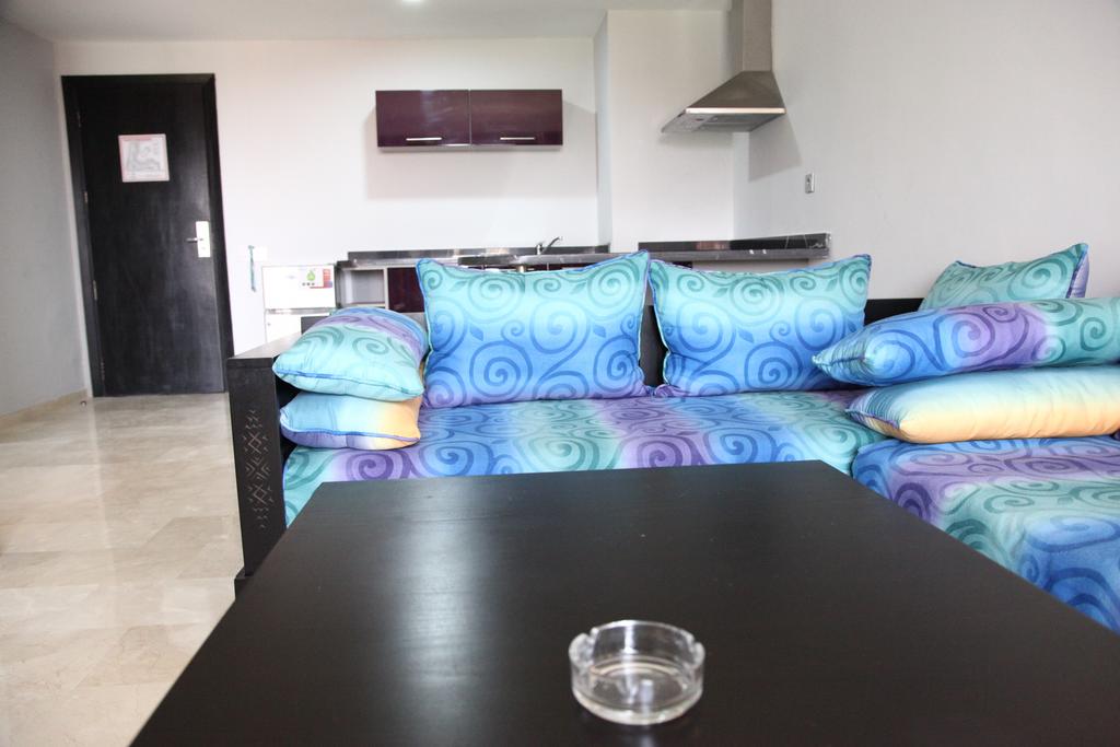 The living area features a sofa adorned with colorful, patterned cushions. A sleek coffee table with a clear ashtray sits in the center. In the background, a kitchenette is visible with modern cabinetry, providing a functional space for meal preparation.