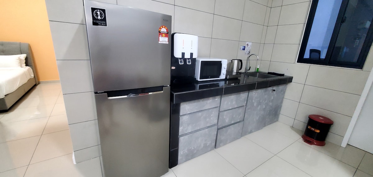 A modern kitchen area features a stainless steel refrigerator, a microwave, and a kettle. The countertop provides ample space for meal preparation. Natural light enters through a nearby window, illuminating the tiled walls and floor.