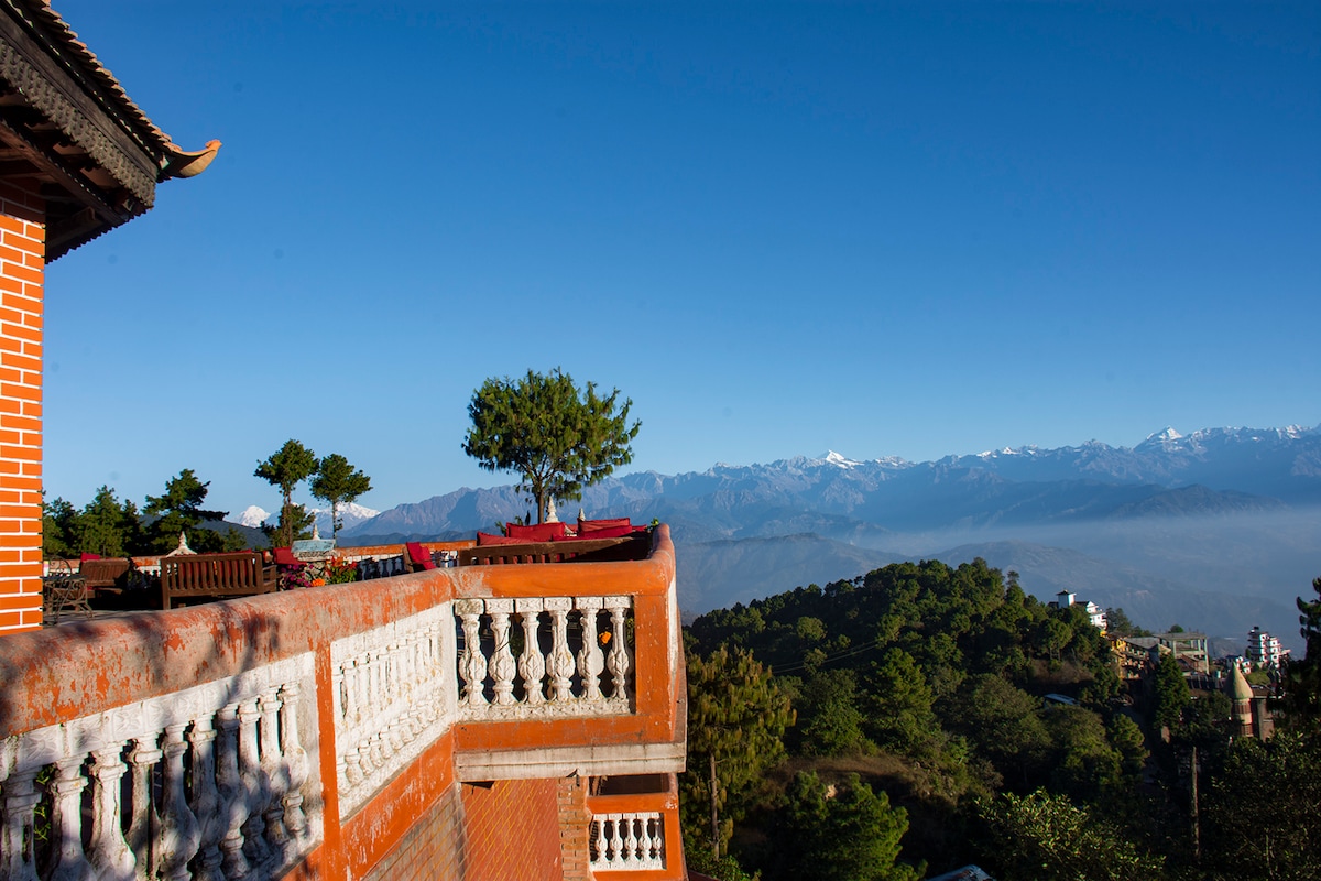 A balcony offers expansive mountain views under a clear blue sky. The railing is adorned with decorative pillars, and a single tree is visible nearby. Seating areas are arranged for relaxation while enjoying the surroundings, highlighting the natural beauty of the location.