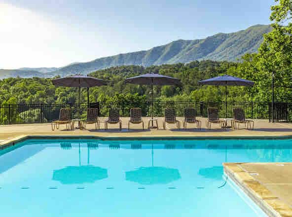 A refreshing outdoor swimming pool reflects the surrounding mountains and blue sky. Several lounge chairs are positioned alongside the pool, each shaded by large umbrellas, creating a relaxing space for guests to unwind amidst nature.