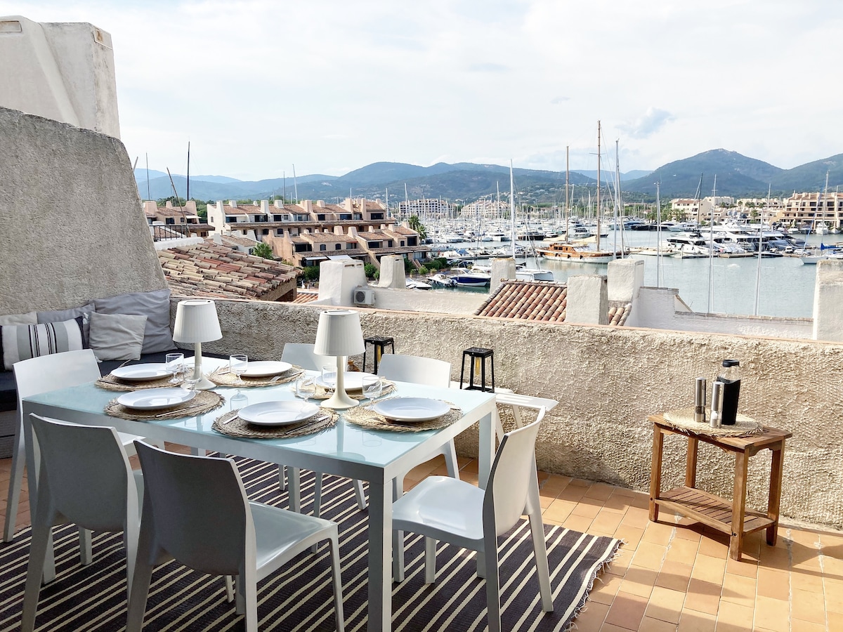 An outdoor dining area is featured with a glass table set for six, surrounded by white chairs. A natural fiber rug lies beneath, and a small wooden side table displays a coffee pot. The marina offers a scenic backdrop with sailboats and distant hills.