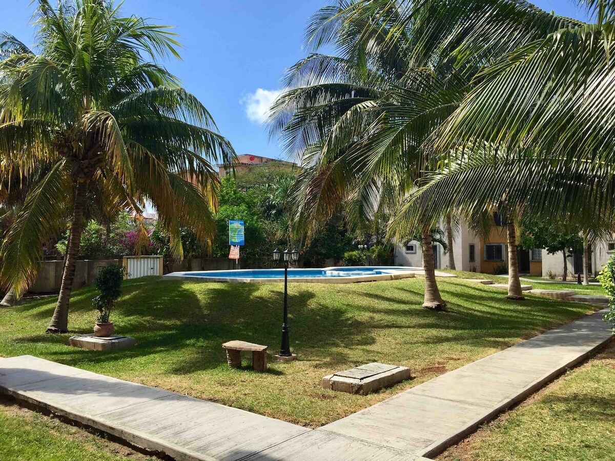 A tranquil outdoor area is shown, featuring well-maintained green lawns bordered by tall palm trees. A pool is visible in the background, surrounded by fresh landscaping. A walkway made of stone connects various parts of the garden, enhancing accessibility and openness.