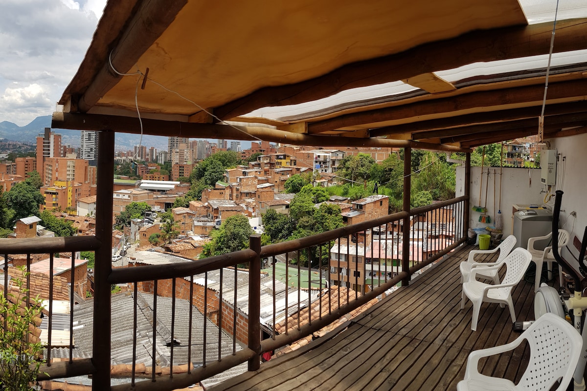 A spacious wooden deck offers views of the surrounding urban landscape, featuring a mix of brick buildings and greenery. Four white chairs are arranged along the railing, inviting relaxation in this outdoor space shaded by a sloped roof, with the city skyline visible in the background.