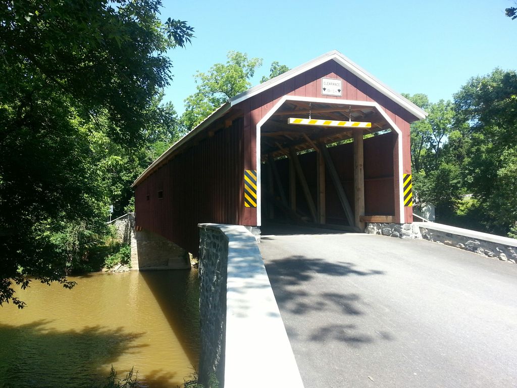 A traditional covered bridge is seen spanning a calm river, supported by wooden beams. The structure features a red exterior with yellow and black caution stripes. Lush green trees surround the area, complementing the serene rural landscape.