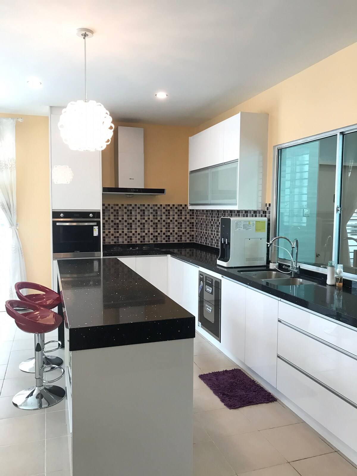 A modern kitchen features a combination of white cabinetry and a black countertop. Bar stools in a vibrant color are positioned at the counter. A window allows natural light to illuminate the space, while a decorative pendant light hangs above for added brightness.