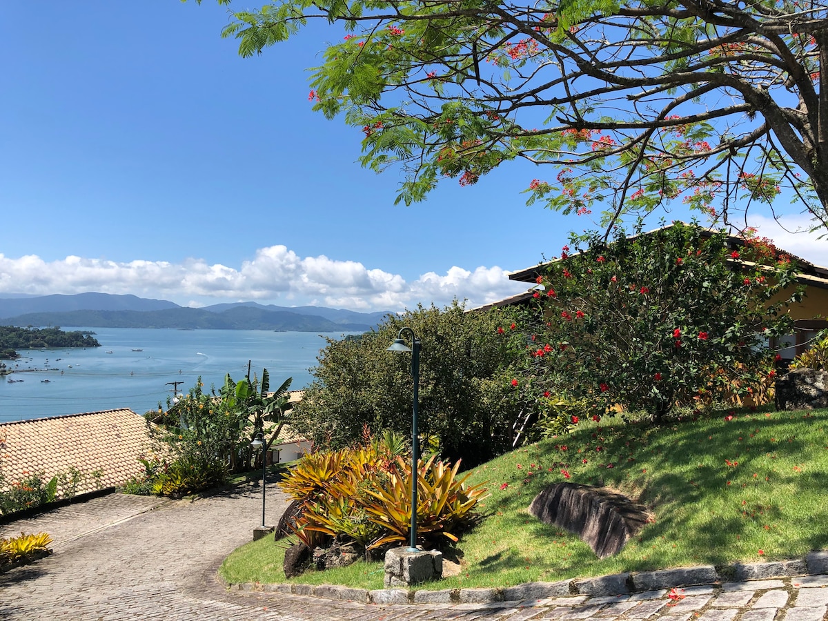 A scenic view captures the waterfront landscape, featuring gentle hills adorned with vibrant greenery and blooming flowers. A winding pathway leads towards a charming yellow house, while the bay and distant islands stretch across the horizon under a clear blue sky.