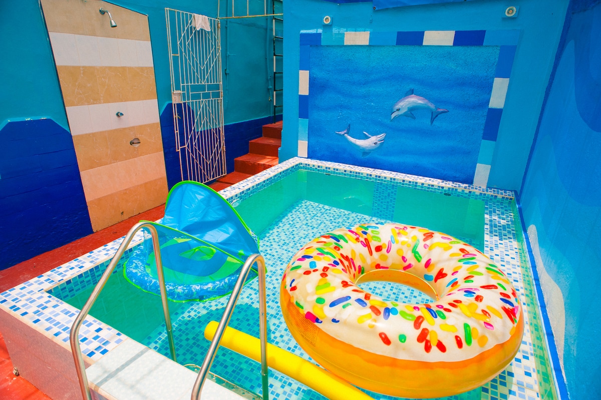 A private pool area is highlighted with a vibrant mural of dolphins. A colorful inflatable ring rests on the water's surface, complemented by a yellow float. A small slide is visible next to the pool, creating an inviting atmosphere for relaxation and fun.