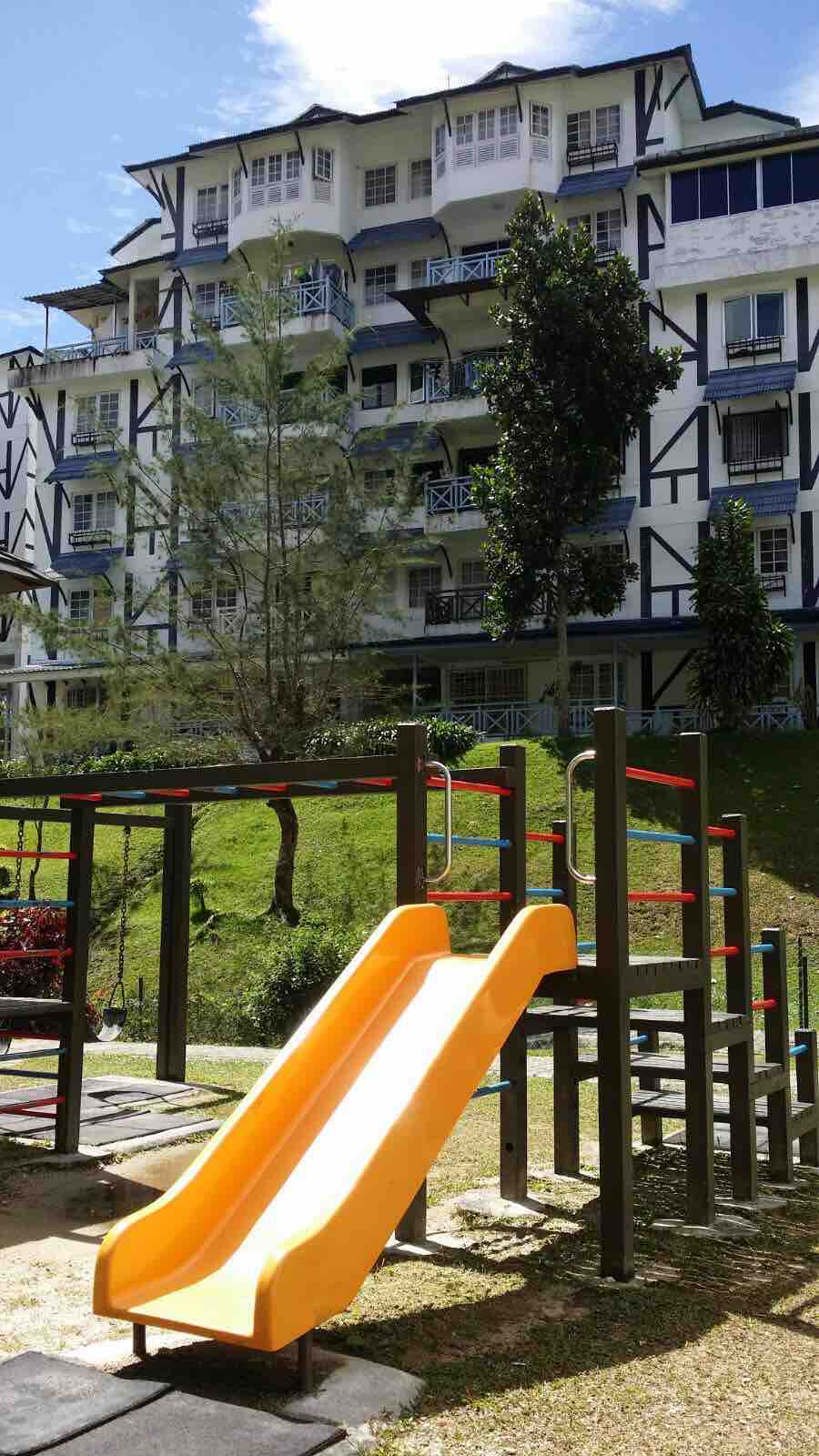 A colorful playground is in the foreground, featuring a bright orange slide and climbing structures. A multi-story building with a traditional design is positioned in the background, surrounded by greenery and trees. The setting offers a family-friendly atmosphere.