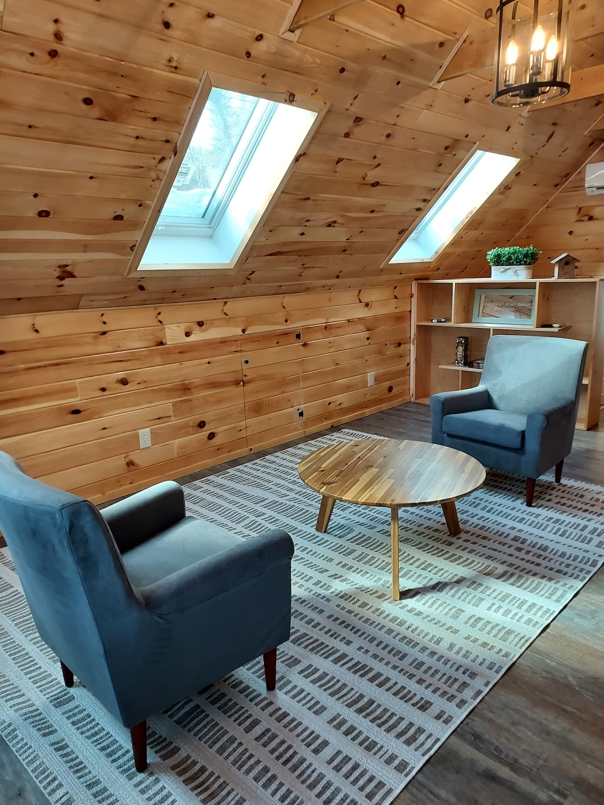 A cozy sitting area features two upholstered chairs and a round wooden coffee table, all set on a patterned rug. Skylights above allow natural light to fill the space, illuminating the warm wooden walls and creating a welcoming atmosphere.