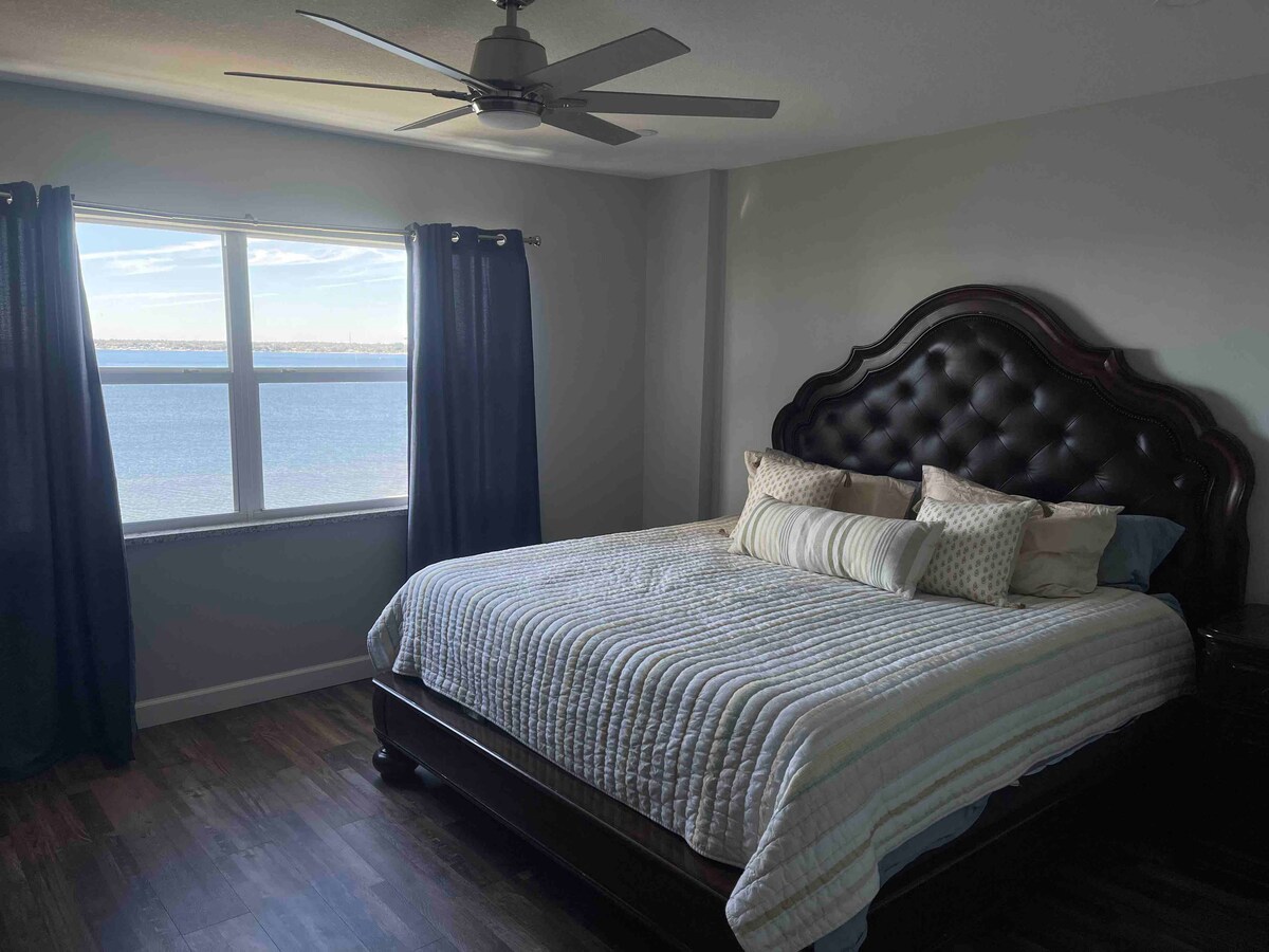 A spacious bedroom is highlighted by a large window offering views of Lake Jackson. A comfortable bed features an elegantly tufted headboard and is adorned with decorative pillows. Dark curtains frame the window, while a ceiling fan adds a modern touch to the room.