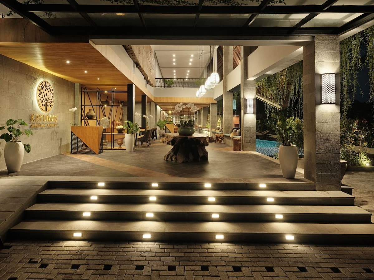 The entrance is highlighted by a series of steps leading up to a welcoming reception area. Soft lighting accentuates the natural stone walls and wooden features. A water feature is visible, adding a tranquil element, while lush greenery surrounds the space, enhancing the overall inviting atmosphere.