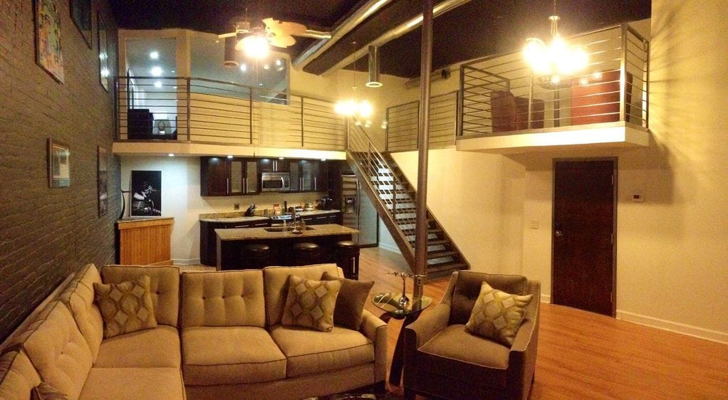 The open layout features a cozy lounge area with a large sectional sofa and two armchairs, complemented by warm wood flooring. An upstairs loft area and a stylish staircase are visible, while the kitchen area showcases a central island and bar seating, illuminated by overhead lighting.