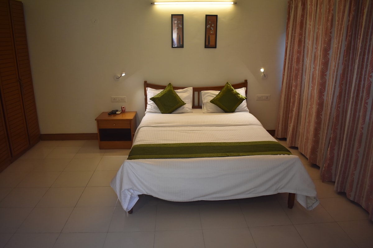 A bright sleeping area features a spacious bed with white linens and green accent pillows. Two framed art pieces adorn the wall above the bed. Soft lighting is provided by wall-mounted fixtures, and a closet stands adjacent to the bed, with light-colored curtains framing a window.