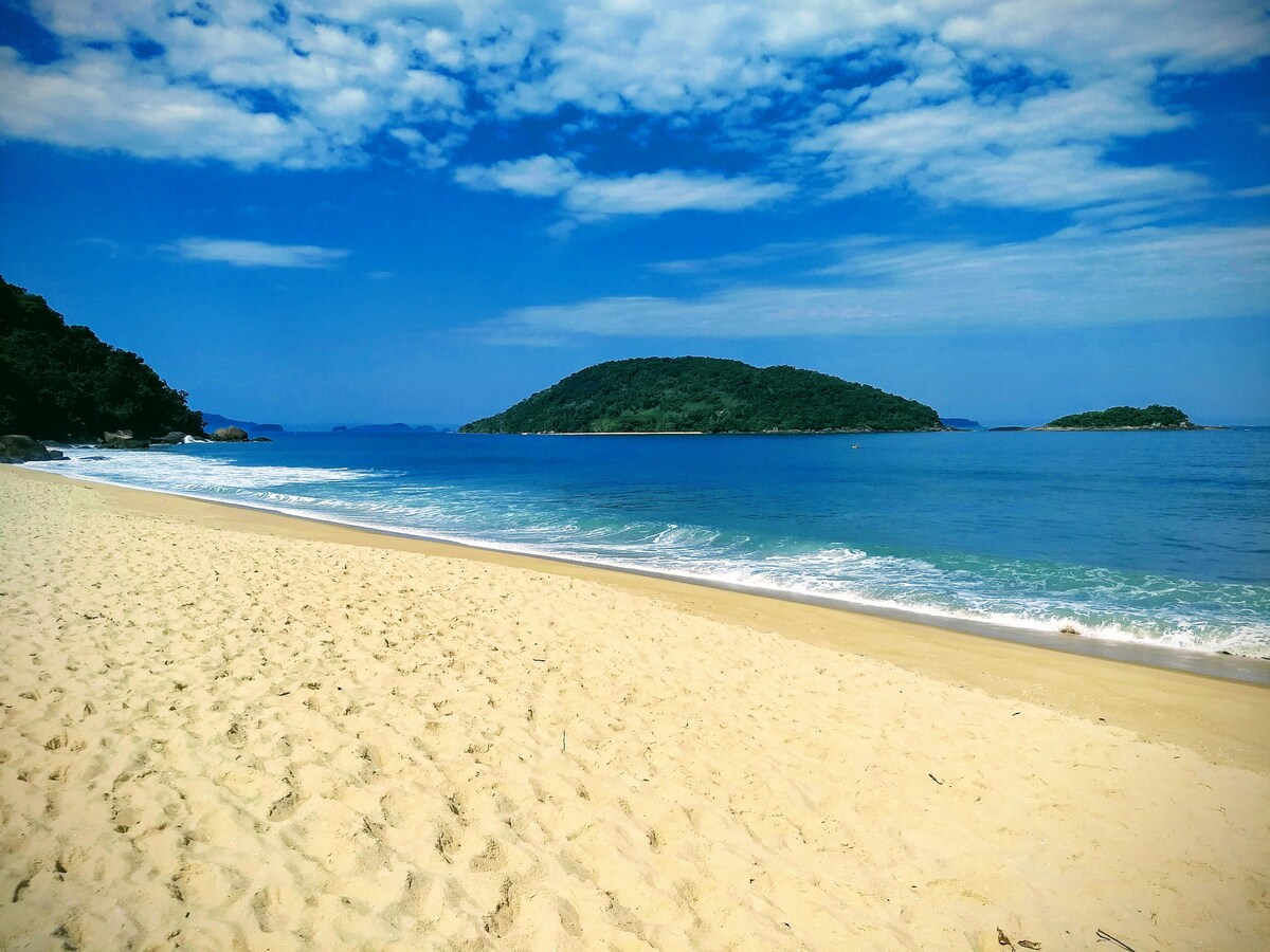 A sandy beach stretches along calm blue waters, with gentle waves lapping at the shore. Lush green islands are visible in the distance, under a bright blue sky dotted with soft clouds, creating a serene coastal landscape.