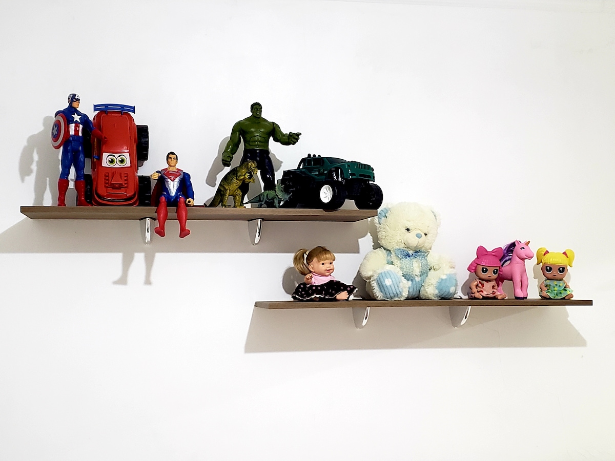 Two wooden shelves display a collection of toys, including action figures, vehicles, and plush animals. The arrangement features colorful characters and soft toys, contributing to a cheerful atmosphere. The wall behind is plain and light-colored, providing contrast to the vibrant toys.