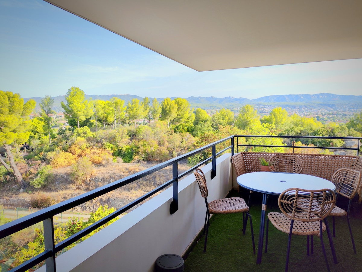 A spacious terrace is presented, furnished with a round table surrounded by several woven chairs. Views of lush greenery and distant mountains can be observed in the background, creating a serene atmosphere for outdoor relaxation.