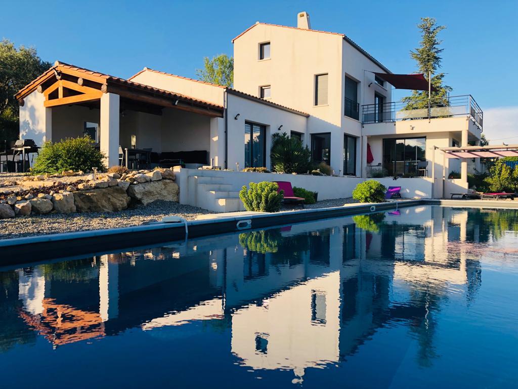 The villa is set against a clear blue sky, featuring a contemporary design with large glass windows. A tranquil swimming pool reflects the building, surrounded by landscaped gardens. Outdoor seating areas are visible, inviting gatherings and relaxation by the water's edge.