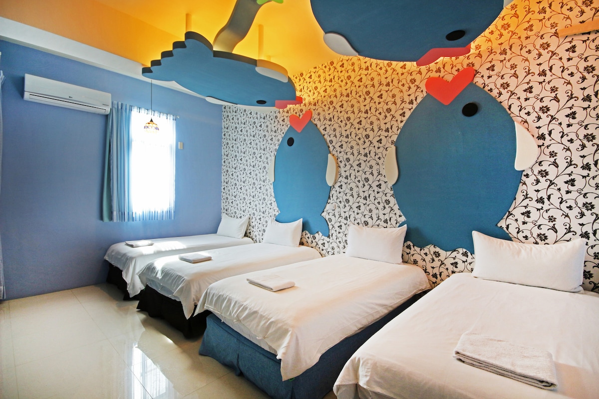 A brightly colored room features several spacious beds, arranged neatly with crisp linens. Playful fish-shaped designs adorn the walls, contributing to the cheerful atmosphere. Natural light filters through a window, complemented by blue drapes, while an air conditioning unit is positioned discreetly above.