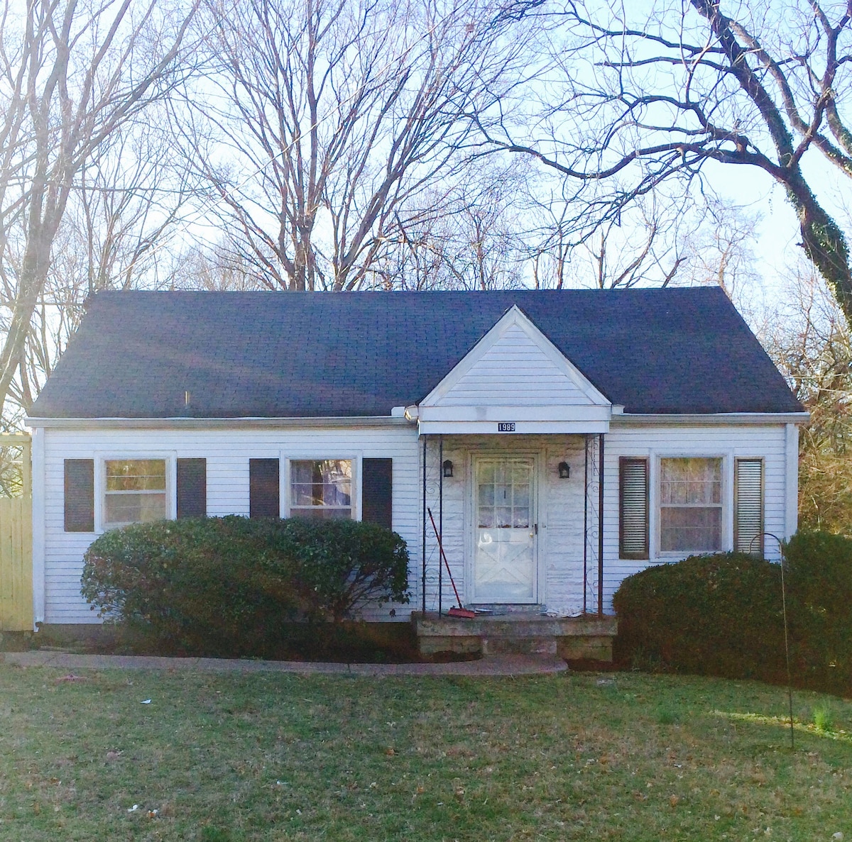 A single-story cottage is set amidst a green lawn, framed by mature trees. The white exterior features black shutters and a welcoming front porch, with a central door flanked by two windows. Neatly trimmed hedges border the space, complementing the clean and inviting appearance.