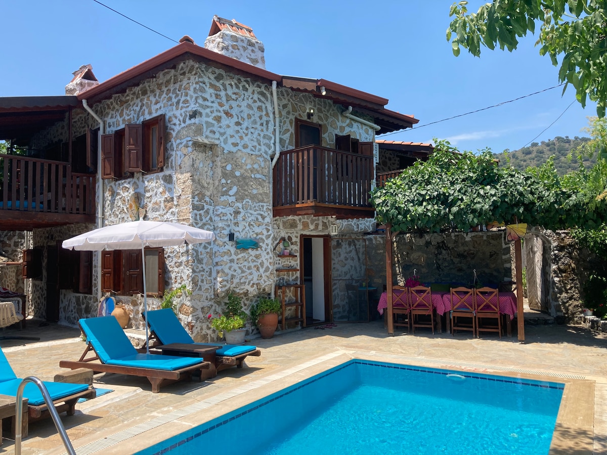 A stone house with a rustic charm is surrounded by greenery and features a sunlit pool. Lounge chairs are positioned beside the water, and a shaded dining area is visible nearby. Balconies on the upper level provide additional outdoor space.