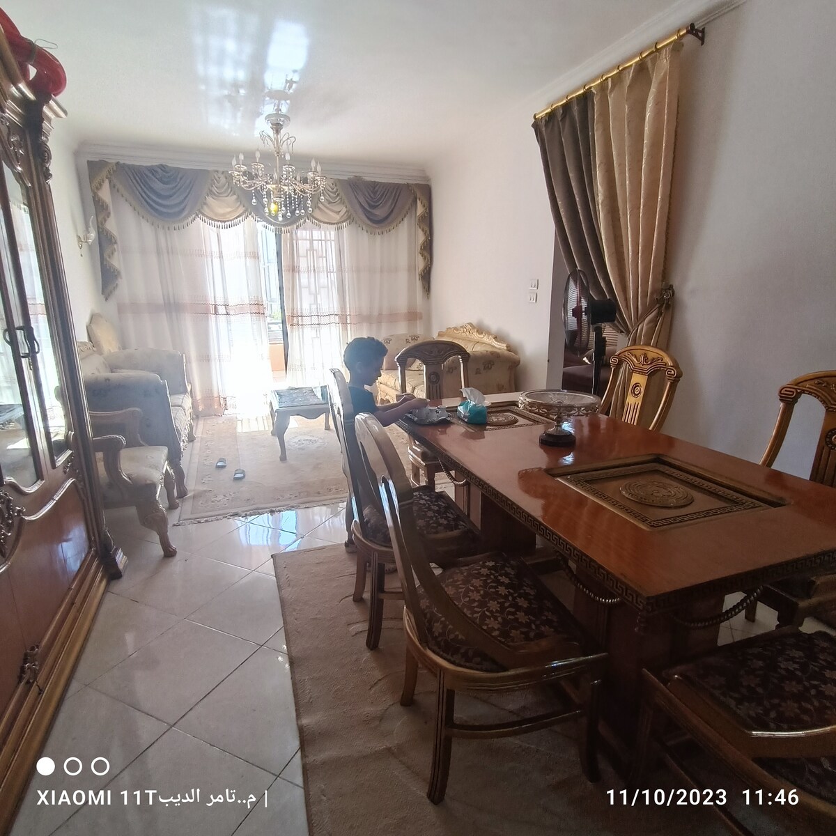 A dining area is presented with a polished wooden table surrounded by several chairs. A decorative chandelier is displayed above, while a cozy seating arrangement with plush sofas is visible in the background. Large curtains frame a window, allowing natural light to enhance the space.
