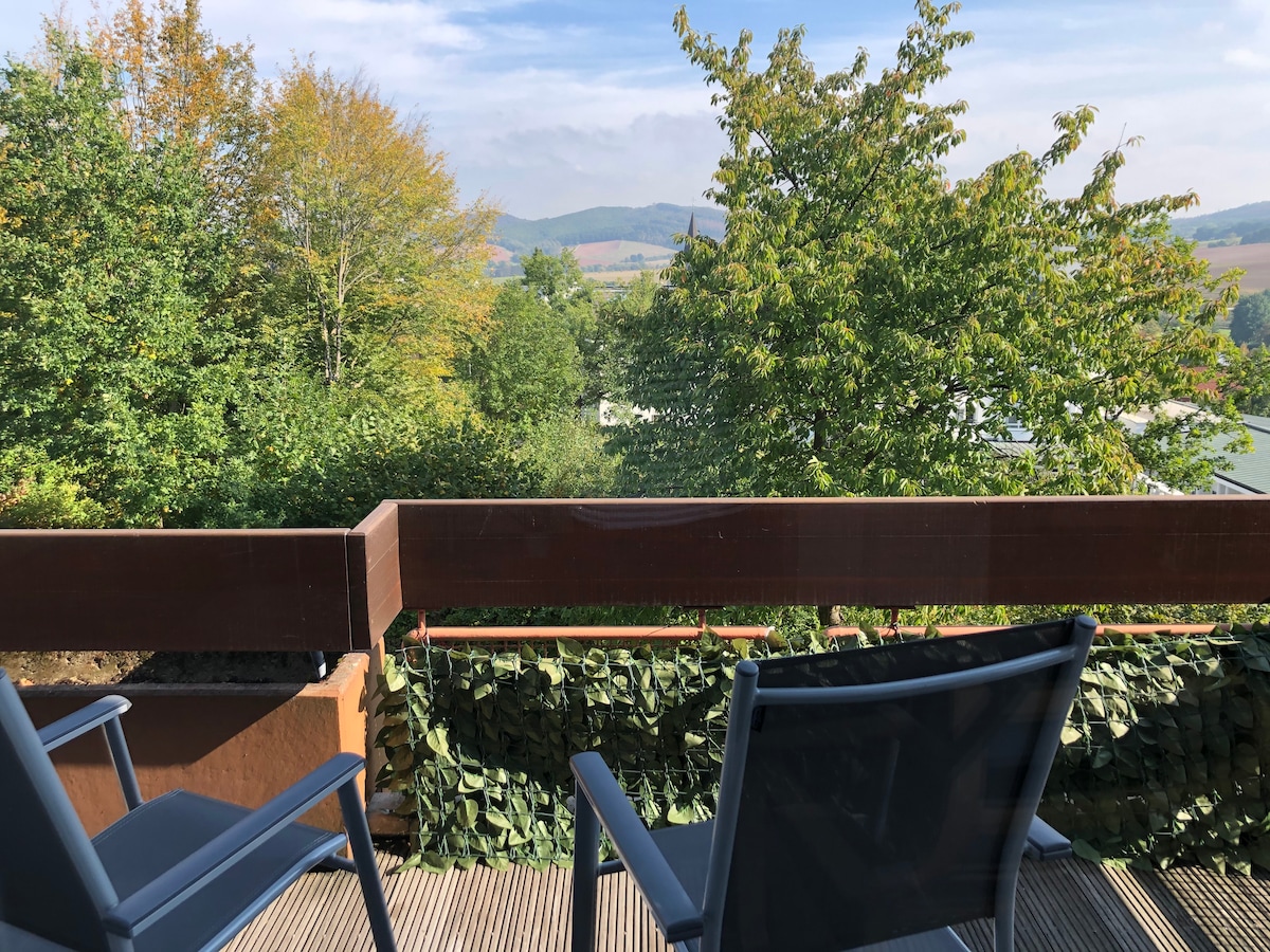 A balcony view reveals lush greenery and distant hills under a clear sky. Two outdoor chairs are positioned on a wooden deck, offering a tranquil space to relax amid the surrounding nature. The railing features decorative greenery, enhancing the sense of privacy.