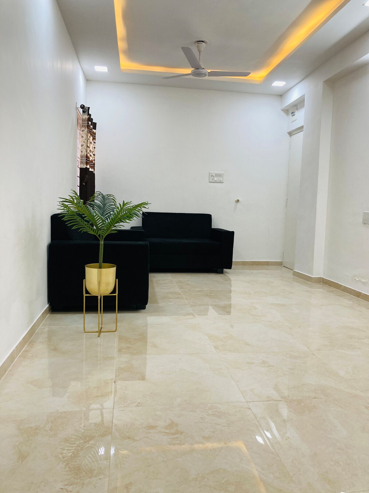 A well-lit living area features a black sofa positioned against a white wall. A small potted plant sits on a gold stand, adding a touch of greenery. The polished tiled floor reflects the soft lighting from the ceiling.