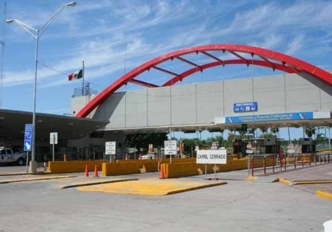 An entrance archway is prominently displayed, marked by a red curved design. A Mexican flag is visible to the left. The area features marked lanes with traffic cones and signage indicating closed passage. Bright blue skies are overhead, enhancing the overall atmosphere.