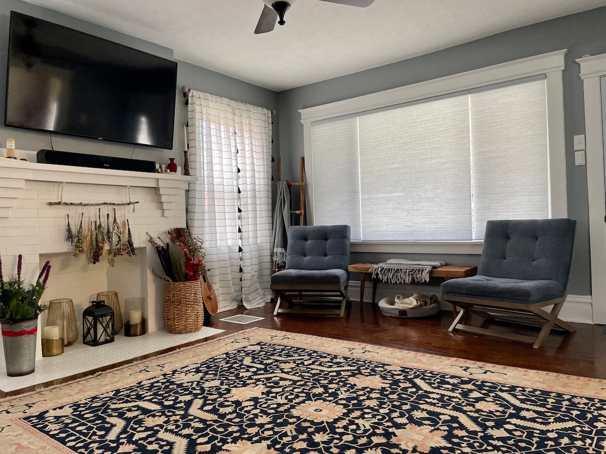 A cozy living area features a modern television mounted above a white-bricked fireplace. Two comfortable chairs are placed across a large area rug, with light filtering through softly covered windows. Decorative plants and a wooden guitar add character to the inviting space.