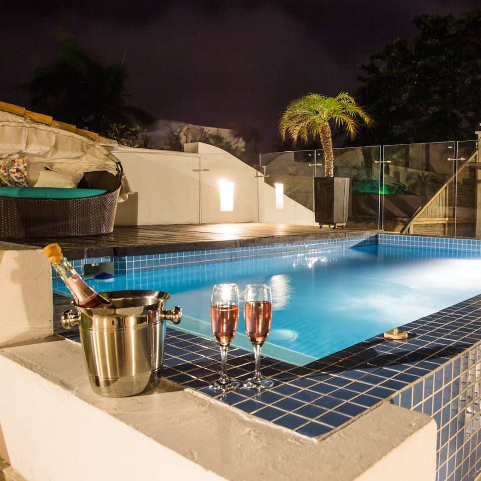 A serene pool area is showcased, featuring a sparkling blue pool surrounded by elegant tile work. An ice bucket with champagne and two glasses sits on the ledge, while palm trees and soft lighting create a relaxed evening ambiance.