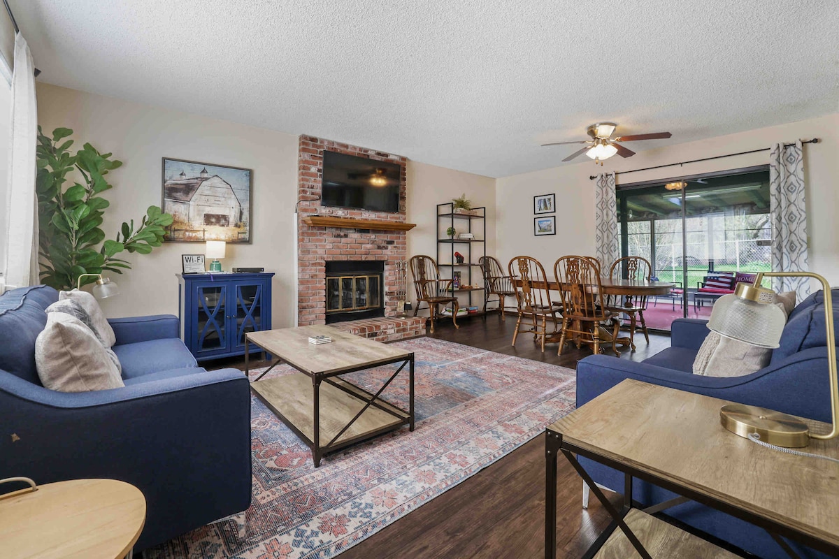 A spacious living room features a comfortable seating area with two blue sofas and a wooden coffee table. A brick fireplace is centered against the wall, with a television mounted above it. A dining area with wooden chairs is visible in the background, enhancing the homey feel.