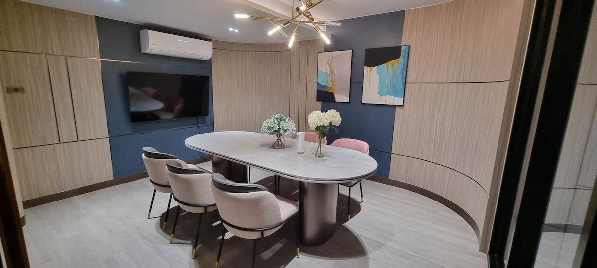 A modern meeting room is presented with a round marble table at its center, surrounded by six soft pink and black chairs. Floral arrangements are displayed on the table, while two abstract art pieces adorn the walls. Natural light softly illuminates the space.
