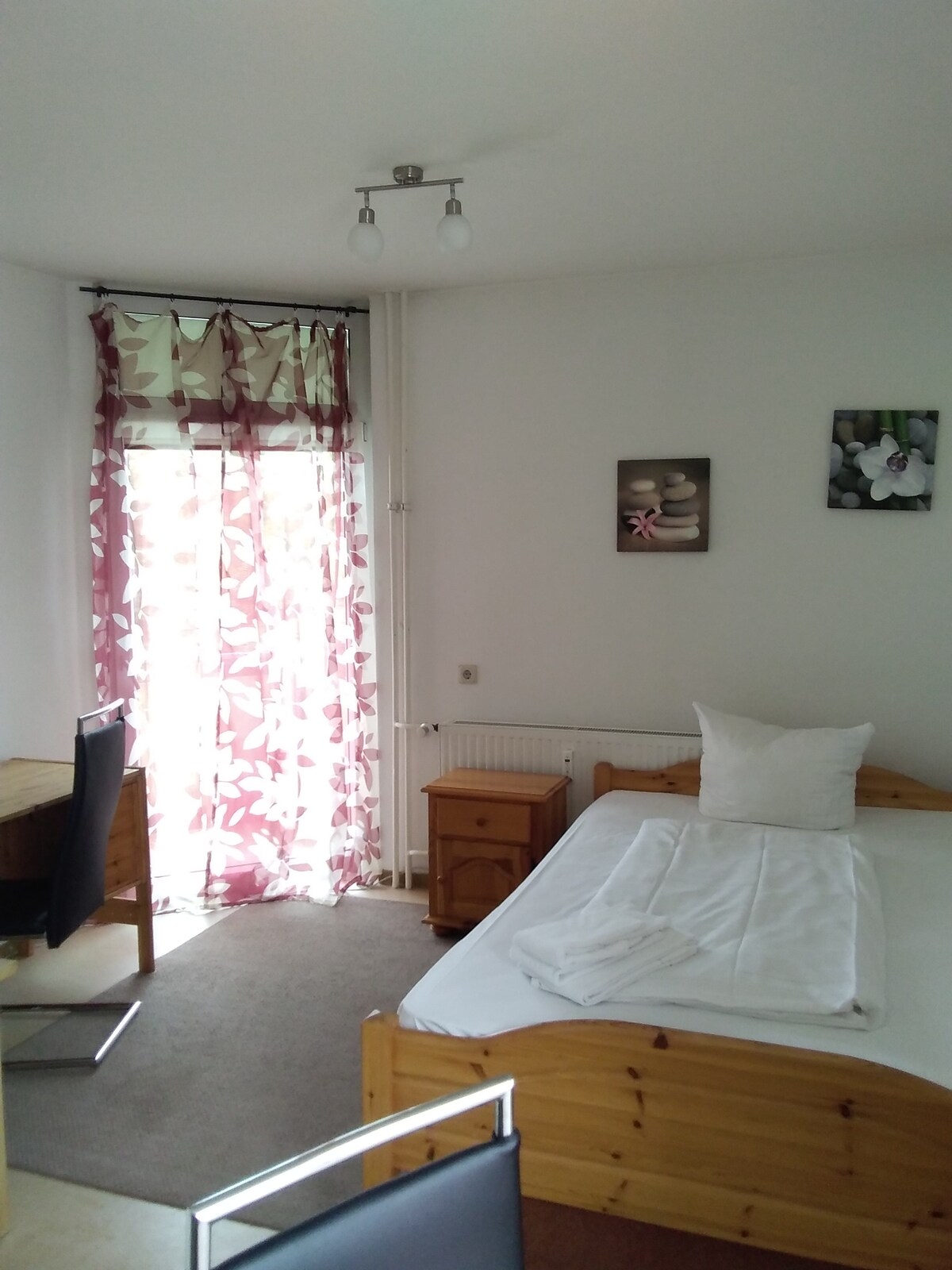 A bright room features a single bed with a neatly arranged white blanket. A small wooden desk and chair are positioned nearby, while a wooden nightstand holds decorative items. Light filters through floral-patterned curtains covering a door that leads to a small outdoor area.