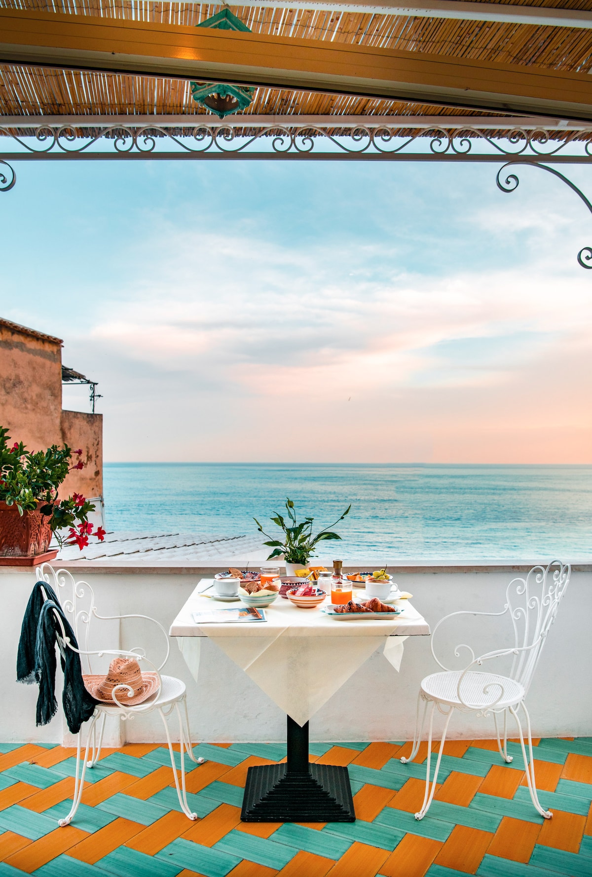 A charming dining area is set on a balcony, featuring a white table adorned with a breakfast spread. Two wrought iron chairs complement the table, while vibrant tiles in shades of orange and turquoise provide a colorful foundation. The ocean is visible in the background under a pastel sky.