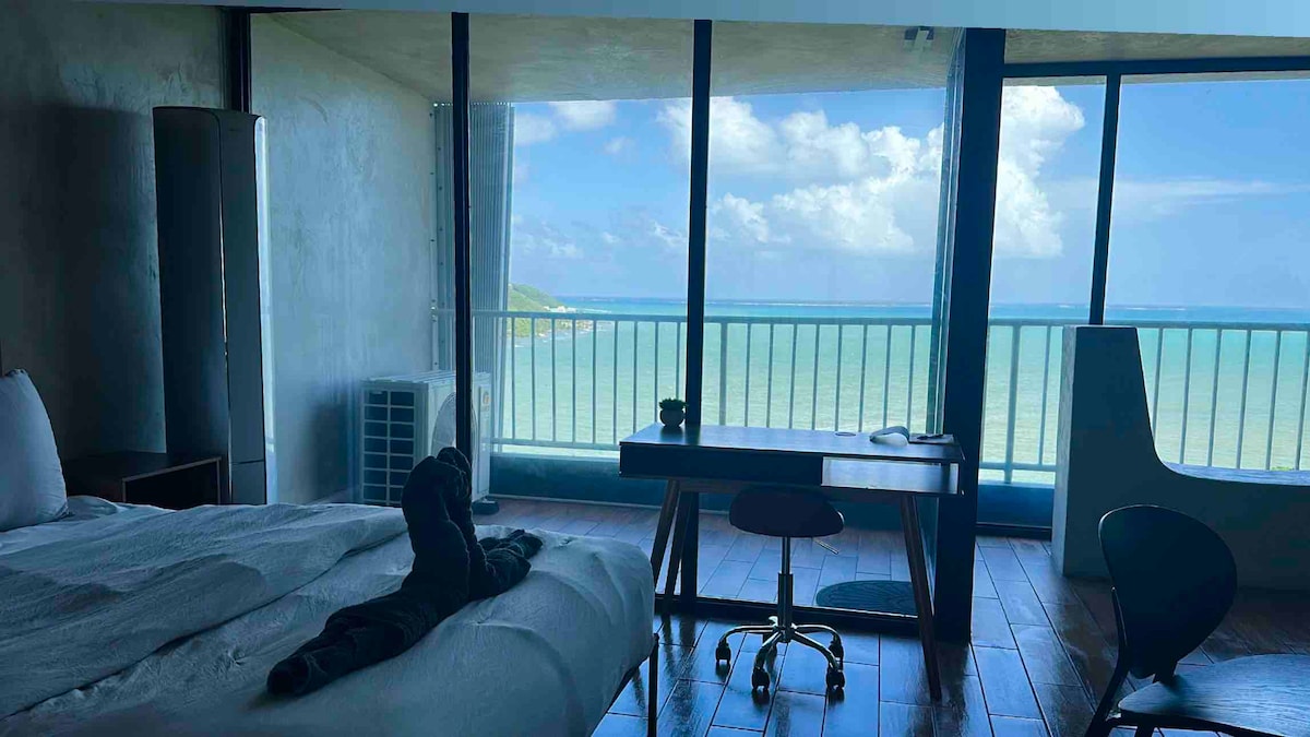 An open-concept studio features a spacious bed with neutral bedding, complemented by a wooden desk and modern seating. Large glass doors offer expansive ocean views, while a cool breeze is suggested through the visible palm-lined coastline. Natural light fills the space, enhancing the serene atmosphere.