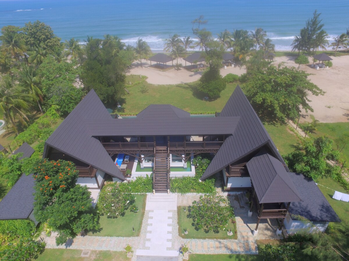 An aerial view of a luxury villa showcases its unique architecture with a distinctive triangular roof. Surrounding gardens provide lush greenery, while the beachfront lies in the background, offering a serene perspective of the ocean and sandy shore.