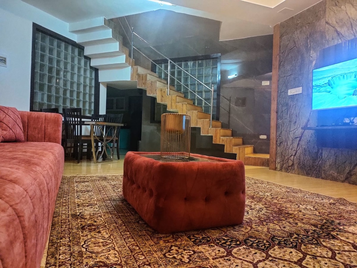 A cozy living area features a plush red sofa and a stylish coffee table placed on a patterned rug. A wooden dining table is visible, accompanied by chairs. A staircase with wooden steps leads to the upper level, providing access to additional living space.